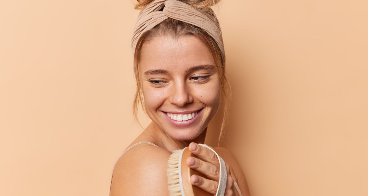 sideways-shot-happy-young-woman-uses-dry-brush-body-has-healthy-smooth-skin-smiles-gently-stands-shirtless-against-beige-background-wears-headband-people-body-treatment-after-shower (1)